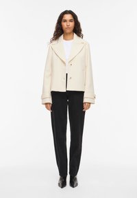 Κρεμ χρώματος κοντό μπουφάν με φαρδιά γιακά, δύο κουμπιά και γυριστά μανίκια, σε συνδυασμό με μαύρο παντελόνι ίσιας γραμμής και μυτερά μποτάκια.