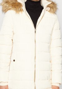 Chaqueta tipo puff en color crema con capucha forrada de pelo, textura acolchada, cremallera frontal y puños acanalados. Detalles en dorado en los bolsillos.