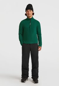 Grön fleecetröja med dragkedja i svart, kombinerad med svarta isolerade byxor och svarta stövlar. Modellen bär en svart mössa.