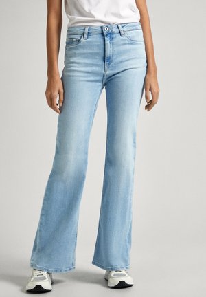 Personne portant un jean flare taille haute bleu clair avec des baskets blanches et un haut blanc, debout devant un fond clair uni.