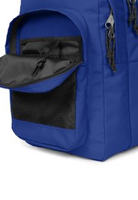 Mochila de tela azul con un bolsillo de malla negra texturizada, que cuenta con un compartimento con cremallera y detalles de hardware en las cremalleras.