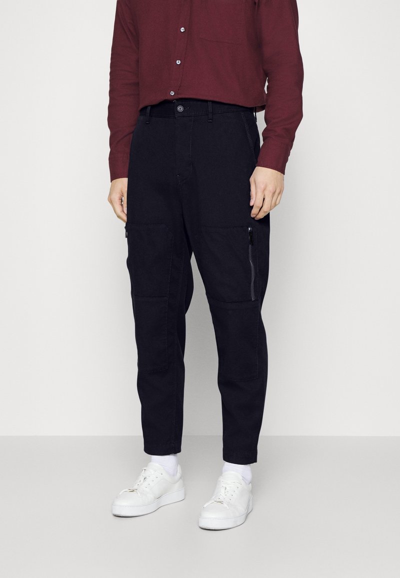 Mand iført mørke cargobukser med lynlåslommer på lårene, en bordeaux skjorte med knapper, hvide sokker og hvide sneakers, stående foran en ensfarvet baggrund.