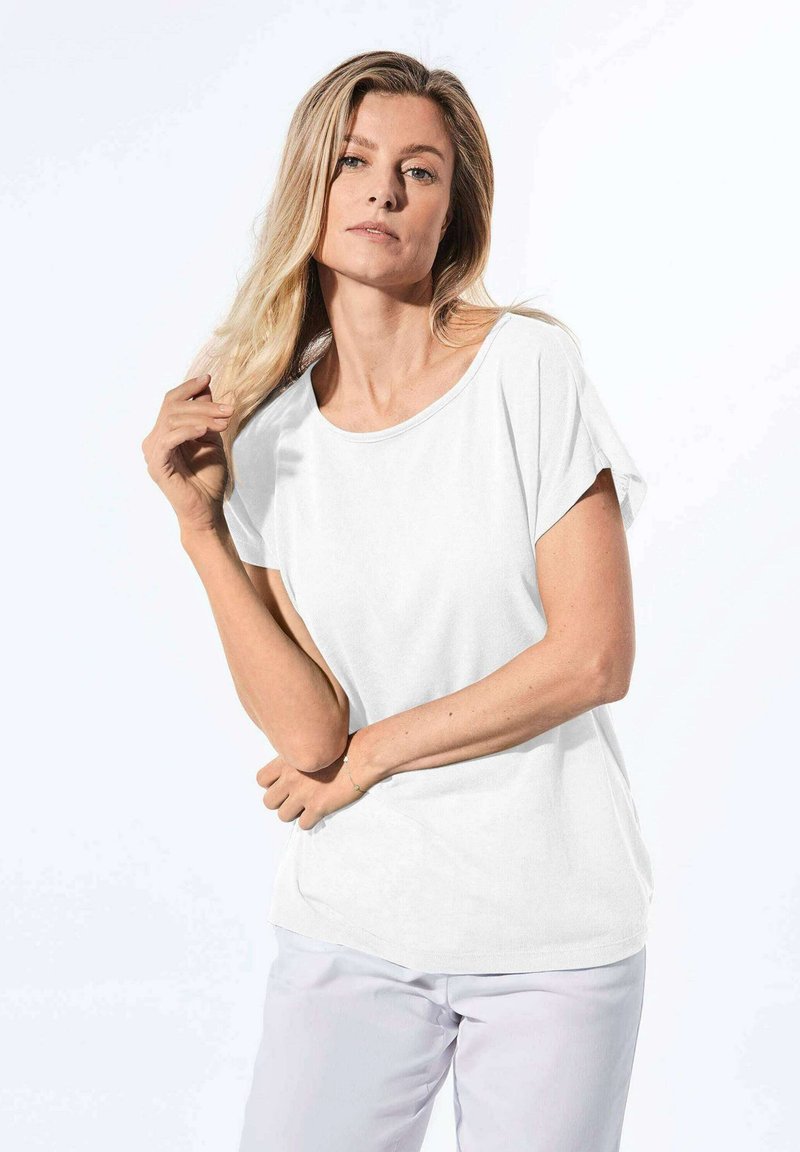 T-shirt bianco a maniche corte con scollatura ampia, texture liscia e vestibilità morbida. Abbinato a pantaloni chiari.
