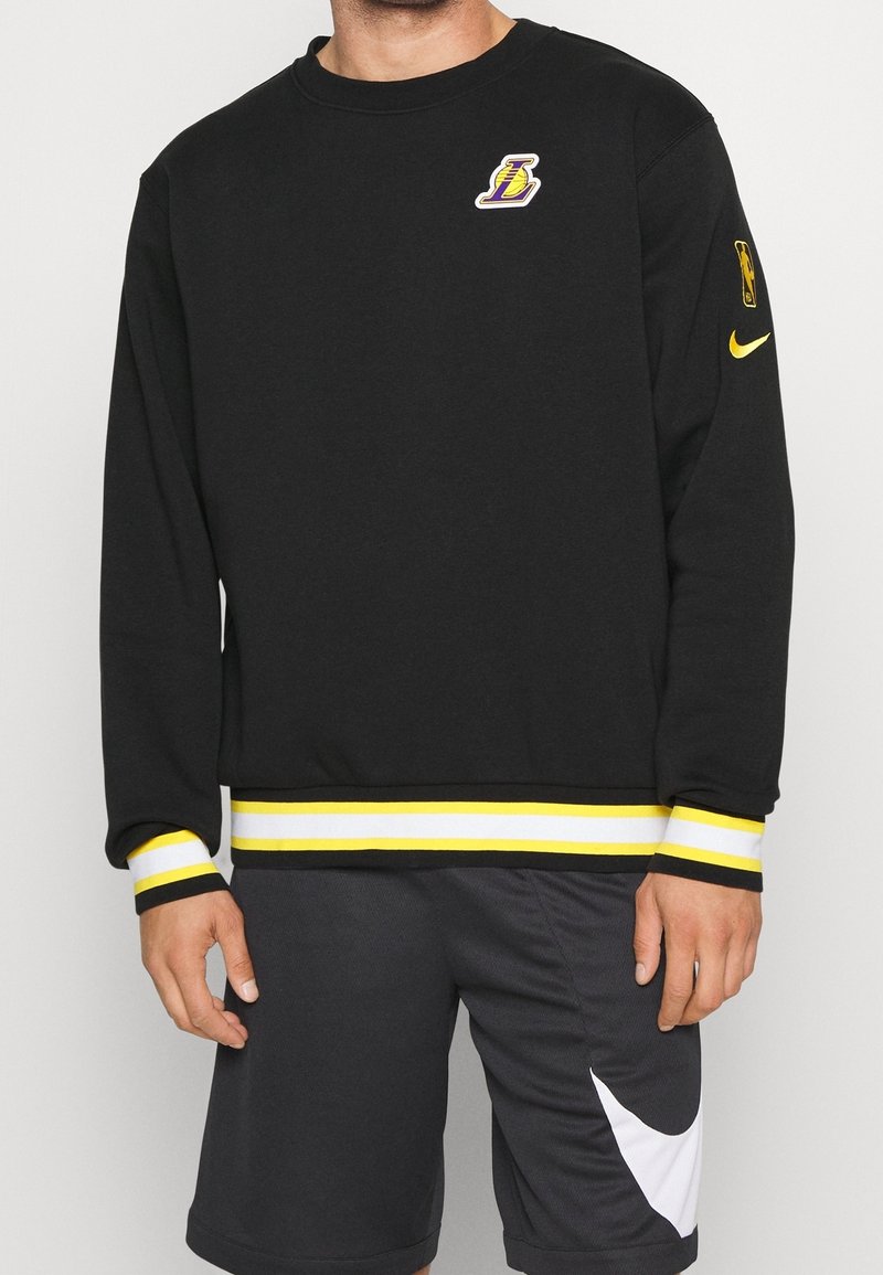 Černá mikina s kulatým výstřihem, bílé a žluté pruhované manžety a spodní lem, na hrudi s fialovým logem Lakers a žlutým swooshem Nike.