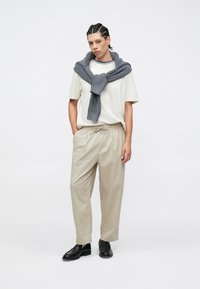 Pantalon ample beige clair avec taille élastique. T-shirt blanc avec col rayé bleu. Pull en maille grise drapé sur les épaules. Chaussures noires.