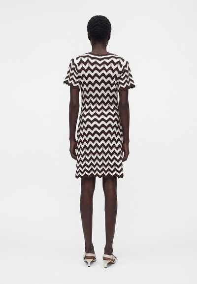 Personne debout de dos portant une robe courte à manches courtes, longueur genoux, avec un motif zigzag noir et blanc et des sandales beiges à petit talon.