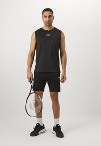 Cămașă sport fără mâneci de culoare neagră cu logo-ul "BOSS", pantaloni scurți negri asortați, ținând o rachetă de tenis și o minge, adidași negri, șosete albe.