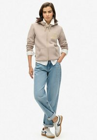 Beige rits-hoodie met koordjes, twee voorzakken, over een witte shirt. Lichtblauwe opgerolde jeans en witte sneakers met zwarte accenten.