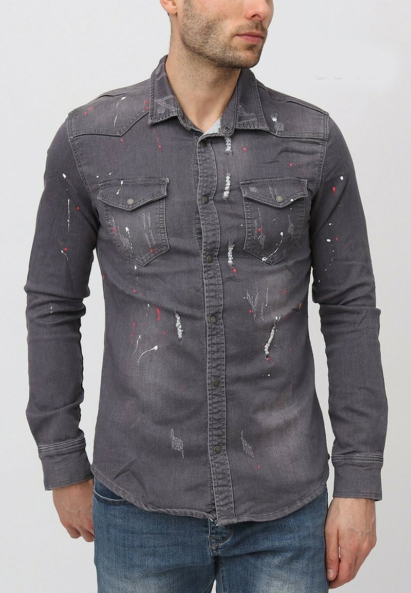 Chemise en denim gris avec un design à boutons, deux poches poitrine et des éclaboussures de peinture blanches et rouges. Manches longues avec poignets.