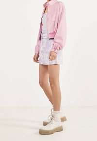 Person som bär en rosa kort jacka, vit linne, lila batikmönstrad minikjol med bälte, vita strumpor och kraftiga vita snörboots.