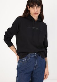 Felpa nera con cappuccio e tasca a canguro, con logo frontale e stringhe. Abbinata a pantaloni in denim blu. Tessuto morbido.