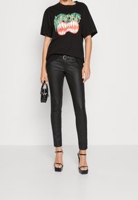 Svart grafisk t-shirt med en färgglad tanddesign, matchad med svarta slim-fit läderbyxor och svarta sandaletter med hög klack och remmar.