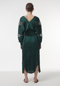 Vestido largo verde plisado con espalda en V y mangas largas. Presenta una cintura ajustada y una tela fluida y texturizada. Se lleva con sandalias negras.