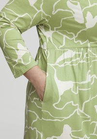 Vestido verde con patrón abstracto blanco, con mangas largas, cintura fruncida y bolsillos laterales. Está hecho de una tela suave y elástica.