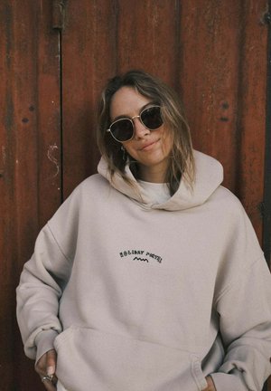 Kobieta o rozpuszczonych brązowych włosach, nosząca okrągłe okulary przeciwsłoneczne i beżową bluzę z kapturem z napisem "Holiday Patrol", stojąca na tle rdzawej czerwonej metalowej drzwi.