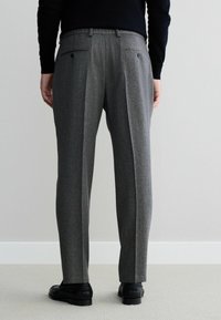 Grau maßgeschneiderte Hose mit Fischgrätenmuster, zwei Gesäßtaschen, eingeknöpftem Frontbereich und elastischem Bund, kombiniert mit schwarzen Schuhen auf einem hellen Boden.