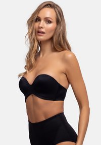 Sort strapless bh med glat, blank overflade og formede skåle, parret med højtaljede sorte trusser, imod en ensfarvet lys baggrund.