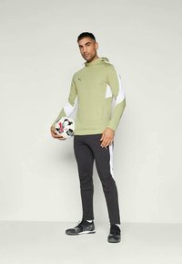 Haut à capuche vert clair avec des accents blancs, pantalon noir ajusté et chaussures de sport noires. Le modèle tient un ballon de soccer noir et blanc.