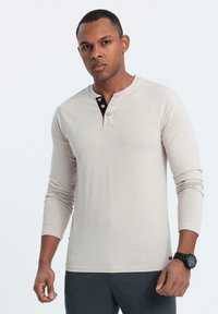 Camicia henley a maniche lunghe color beige chiaro con colletto rotondo, pattina nera con bottoni e tre bottoni marroni. Materiale in cotone, vestibilità slim.