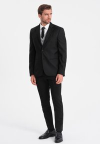 Musta puku tarkasti istuvana, valkoinen paita, musta solmio ja harmaa liivi. Mustat kengät, joissa on sileä nahkapinta ja minimalistinen muotoilu.