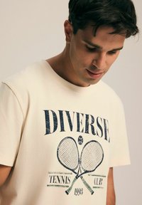 Kremowa bawełniana koszulka zniszczona w granatowym nadruku przedstawiającym rakiety tenisowe oraz tekst "DIVERSE TENNIS CLUB 1993" w retro wzorze.