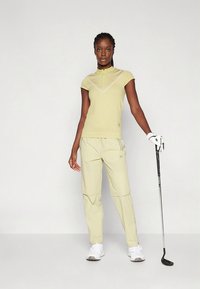 Jasnożółta koszulka polo z krótkim rękawem i suwakiem przy szyi; w zestawie jasnożółte spodnie z kieszeniami. Model trzyma kij golfowy, ma na sobie białe buty.