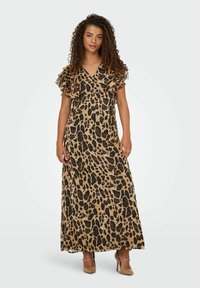 Donna con capelli ricci che indossa un lungo vestito con stampa leopardata, con maniche corte a volant e scarpe con tacco beige, in piedi su uno sfondo neutro.