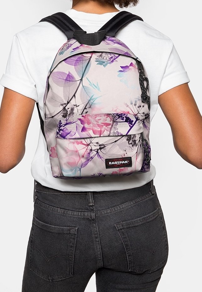 Personne portant un sac à dos Eastpak à motif floral avec des fleurs roses, violettes et bleues sur une chemise blanche et un jean foncé.