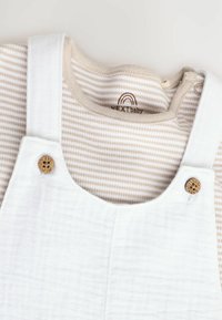Weiße Baumwolloveralls mit Holzknopfakzenten, über einem beigen und weißen gestreiften Langarmshirt mit rundem Kragen kombiniert.