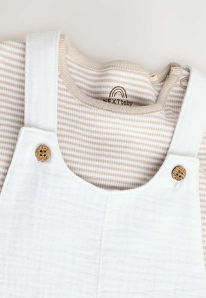 Baltas kokvilnas kombinezons ar koka pogu akcentiem, uzvilkts pār gaiši brūnu un baltu svītrainu garo piedurkņu krekli ar apaļu dekoltē.