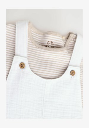 Baltas kokvilnas kombinezons ar koka pogu akcentiem, uzvilkts pār gaiši brūnu un baltu svītrainu garo piedurkņu krekli ar apaļu dekoltē.