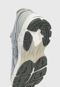 Semelle de chaussure de course grise et blanche avec des patins d'adhérence noirs et un motif de bande de roulement détaillé incliné vers le haut sur un fond uni.