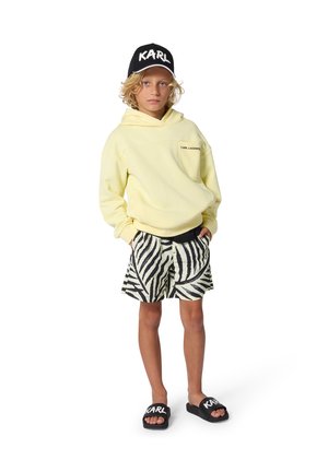 Sweat à capuche jaune avec logo sur la poche avant, short à rayures zébrées noir et blanc, casquette noire avec "KARL" et sandales slides noires.