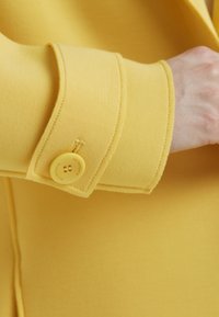 Gelber Jackenärmel mit einem passenden gelben Knopf mit eingravierten Details. Der Stoff hat eine glatte Textur und ein maßgeschneidertes Design.