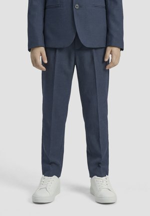 Person wearing navy blue dress pants, matching blazer, and white sneakers, standing against a plain light background.