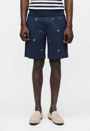 Hombre que lleva pantalones cortos azul marino con pequeñas banderas bordadas multicolores, combinados con mocasines beige y una camisa azul marino y blanca a rayas.