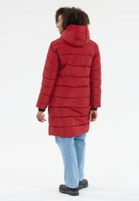 Röd vadderad lång jacka med huva, med horisontella quiltade sektioner, långa ärmar och elastiska mudd. Den är dragen över blå denimjeans.