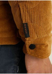 Gros plan d'une manche de veste en velours côtelé marron avec un bouton noir et une étiquette en tissu noire portant la mention "CHASIN'".