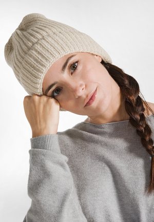 Lichtbeige geribbelde gebreide beanie met een relaxed fit. Zachte textuur en een slouchy ontwerp aan de bovenkant. Draagt bij een grijze, loszittende trui.