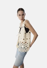 Chaleco de ganchillo en color crema con cortes circulares y flecos decorativos, combinado con una blusa negra y pantalones cortos de mezclilla azul claro.