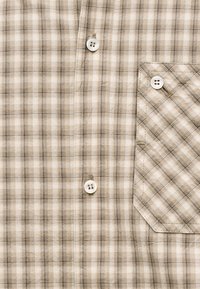 Camisa de cuadros beige y marrón vista frontal, con tres botones blancos y un bolsillo en el pecho a juego con botones en el lado derecho.