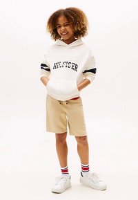 Hvit hettegenser med marineblå striper og "HILFIGER"-tekst, beige shorts, hvite sneakers og sokker med rød-hvite-blå striper. Krøllete hår.