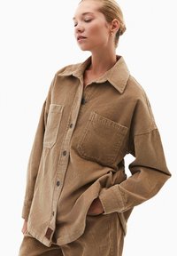 Camicia in corduroy marrone con collo, due tasche frontali, chiusura a bottoni e vestibilità ampia, caratterizzata da una texture a coste verticali.