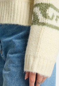 Suéter de punto crema con puños acanalados, que presenta un patrón geométrico verde a lo largo del pecho. Combinado con jeans de mezclilla azul.