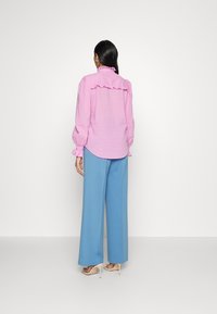 Pink textured blouse with ruffled collar and sleeves, paired with wide blue trousers, set against a plain white background.