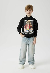 Un sweat à capuche noir avec un imprimé graphique représentant un personnage et un texte, associé à un jean ample bleu clair et des baskets blanches.