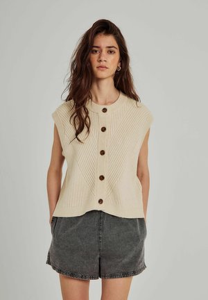 Beige ujjatlan kötött felső, függőleges bordázással, öt barna gombbal az elején, és kerek nyakkivágással. Szürke rövidnadrággal párosítva.