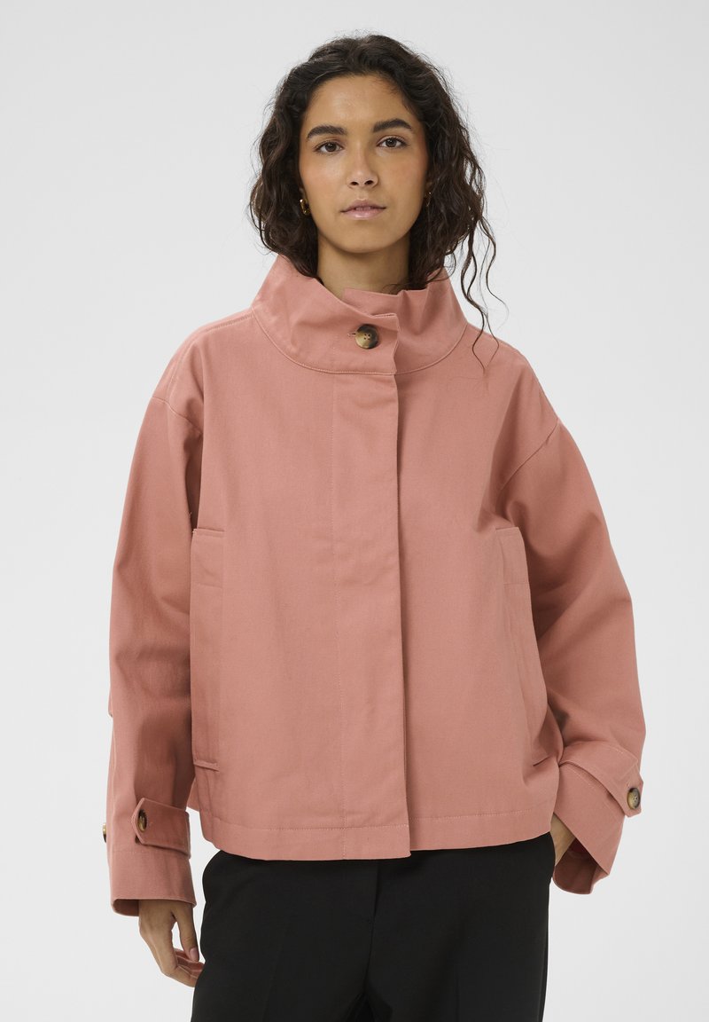 Kohvri juustega naine, kes kannab lõhesinine-roosat nupukestega jopet ja musti pükse, seisab ühevärvilise heleda tausta ees.