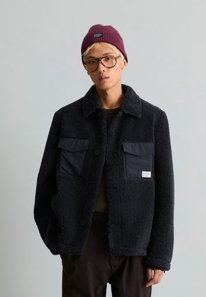 Jeune personne aux cheveux clairs porte de grandes lunettes, un bonnet bordeaux, une veste noire texturée avec des poches avant, debout devant un fond gris uni.
