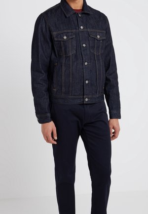 Hombre con chaqueta vaquera azul oscuro con botones metálicos y pantalones oscuros, de pie frente a un fondo neutro.